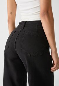 Zwarte high-waisted jeans met een relaxed fit, voorzien van twee achterzakken en zichtbare stiksels. De stof heeft een gladde, licht gestructureerde afwerking.