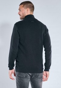 Schwarzer Pullover mit strukturierter Rückseite, glatten Ärmeln, hohem Kragen und einem kleinen Logodetail. Kombiniert mit grauen Jeans, Modell blickt von der Kamera weg.