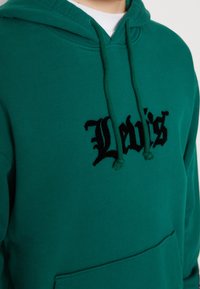 Grüne Hoodie aus Baumwolle mit einer vorderen Kängurutasche, versehen mit schwarzem, gesticktem "Levi's"-Text und passenden Kordeln.