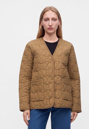 Femme aux cheveux blonds raides portant une veste matelassée marron avec des boutons-pression et deux poches avant, associée à un jean bleu et un haut noir.