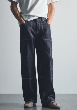 Personne portant un pantalon en jean foncé ample avec des coutures visibles, les mains dans les poches, associé à une chemise blanche et des baskets beiges sur un sol gris.