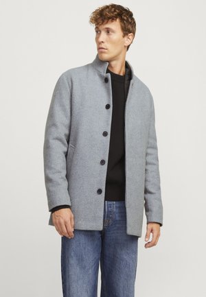 Abrigo de lana gris con cuello alzado, cierre frontal con botones y bolsillos laterales, llevado sobre un suéter negro y jeans azules.