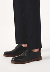 Črni usnjeni čevlji z zaobljenim prstom, vezalkami in rjavim gumijastim podplatom. Kombinirani s temnimi, širokimi hlačami.