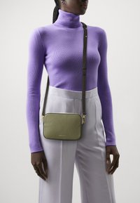 Borsa crossbody verde con texture, di forma rettangolare, con hardware dorato e tracolla marrone, indossata sopra un dolcevita viola e pantaloni chiari.
