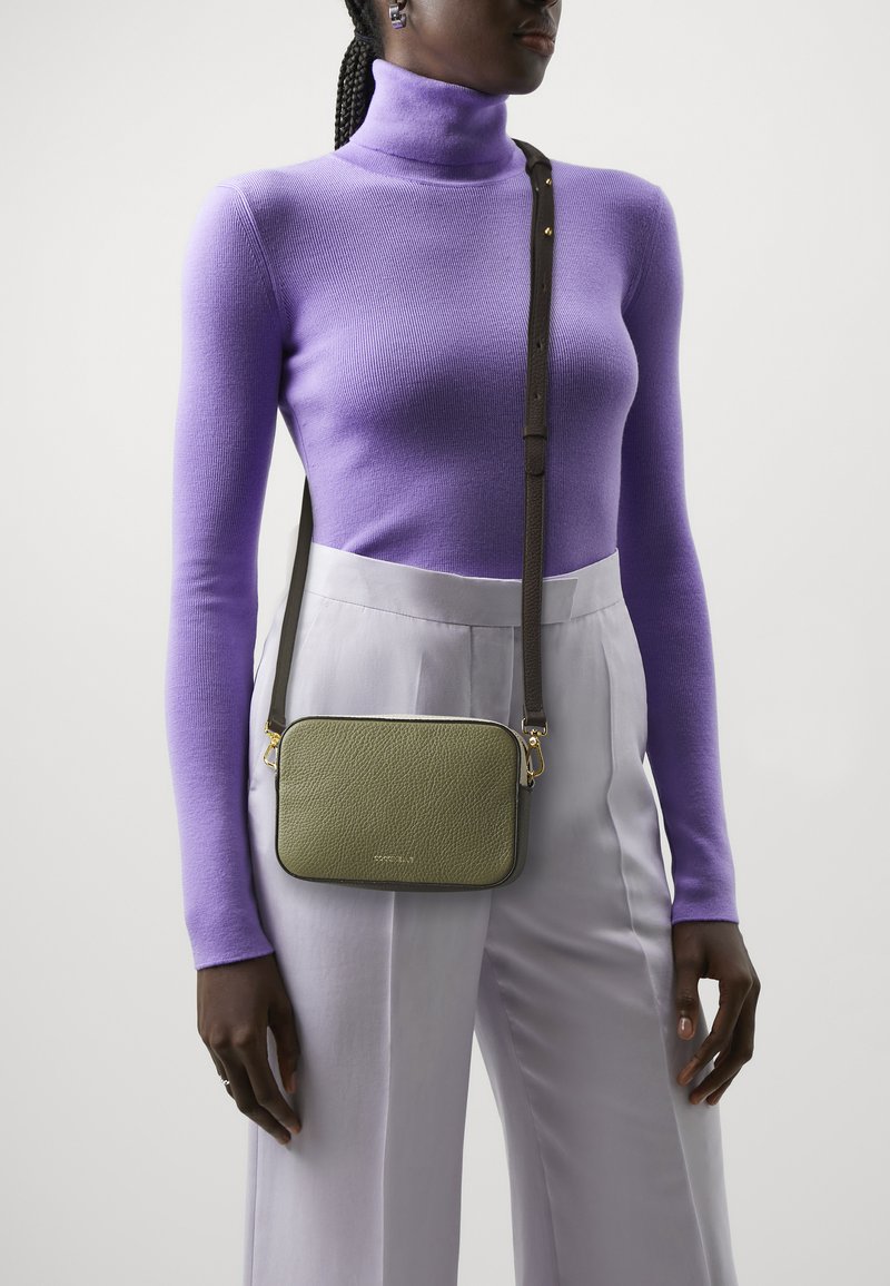 Borsa crossbody verde con texture, di forma rettangolare, con hardware dorato e tracolla marrone, indossata sopra un dolcevita viola e pantaloni chiari.