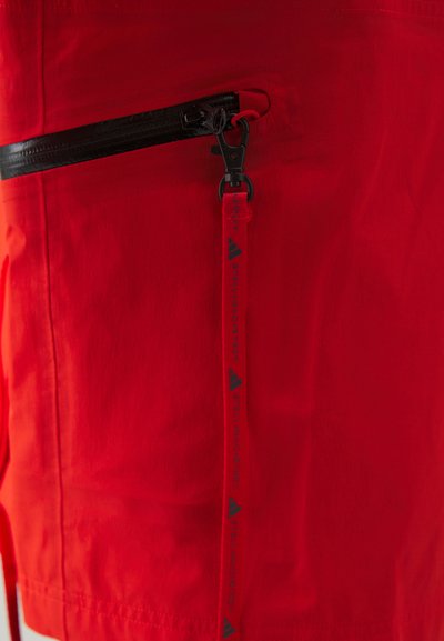 Red fabric garment featuring a black zippered pocket, textured finish, and a contrasting branded pull tab with black text.