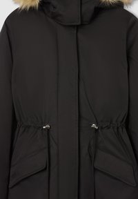 Czarna parka z kapturem wykończonym futrem, ze sznurkiem w talii i dwoma przednimi kieszeniami z klapami. Gładki materiał o matowym wykończeniu i detalami stębnowania.