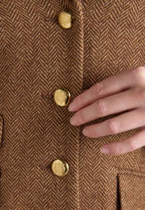 Tissu marron en chevrons avec trois boutons dorés ornés de blasons en relief, et une main aux ongles naturels touchant le bouton du milieu.