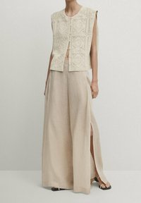 Ärmlös beige virkad väst i kombination med breda, högmidjade beiga byxor med sidslitsar och svarta sandaletter med öppen tå.