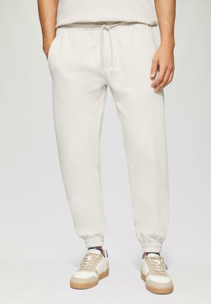 Pantalones de chándal gris claro con cintura y puños elásticos, que cuentan con bolsillos laterales y una textura de tela suave y lisa. Llevados con zapatillas beige.