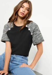 Sort T-shirt med ternede tweedærmer, der har sølv pyntede knapper og frynsekant. Model iført blå jeans.
