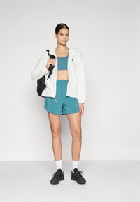 Lichte witte zip-up hoodie, teal sportbh en bijpassende shorts. Zwarte rugzak en sneakers. Eenvoudig ontwerp, gericht op comfort.