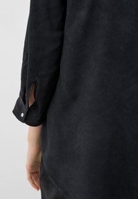Braccio sinistro di una persona in una camicia nera a maniche lunghe con bottone al polsino e una piccola fessura vicino al polso, su uno sfondo chiaro e uniforme.