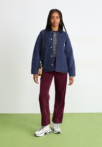 Giacca di tela blu navy con tasche frontali sopra una camicia a righe, abbinata a pantaloni in velluto a coste bordeaux e sneakers bianche con dettagli neri.