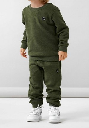 Enfant portant un sweat-shirt vert olive et un pantalon de jogging assorti avec des baskets blanches, debout devant un fond clair uni.