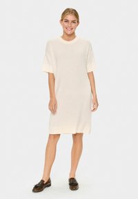 Femme debout et souriante, portant une robe en tricot crème jusqu'aux genoux avec des manches courtes et des mocassins marron.