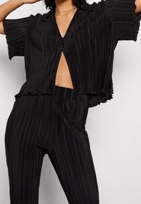 Zwarte geplooide top met korte mouwen en een diepe V-hals, met een knoopsluiting en een gerimpelde zoom, gecombineerd met bijpassende geplooide broek.