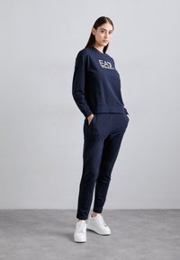 Tumši zils kapučīns un atbilstoši joggeri, kas izgatavoti no mīksta auduma, ar zeltītu logo uz krūšu daļas, ar sānu kabatām un brīvu piegriezumu.