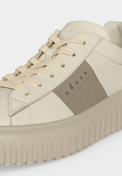 Gros plan d'une basket beige avec une semelle épaisse, des lacets assortis et un panneau latéral gris affichant le texte "HØN86" et un petit symbole d'aile.