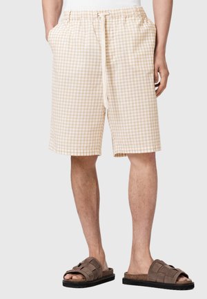 Person trägt beige-weiß karierte Shorts mit Kordelzug und Seitentaschen sowie braune Sandalen mit offenen Zehen, steht vor einem schlichten Hintergrund.