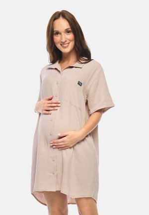 Schwangere Frau in locker sitzendem, beigem Knopfkleid hält sanft ihren Bauch und lächelt vor schlichtem weißem Hintergrund.