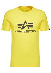 Žluté bavlněné tričko s výrazným čern�ým logem s křídly a textem: "ALPHA INDUSTRIES KNOXVILLE, TENNESSEE, U.S.A." na přední straně.