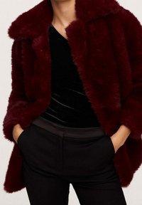 Mujer con un abrigo de piel sintética de color rojo oscuro sobre una blusa negra y pantalones negros, con las manos en los bolsillos, torso y parte superior de las piernas visibles.