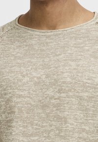 Close-up of a beige and light brown marled knit sweater worn on a person’s upper chest and neck area.