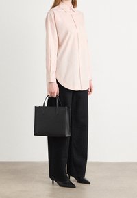 Person som bär en ljusrosa knäppt skjorta, svarta vida byxor, svarta spetsiga klackskor och håller en strukturerad svart handväska i handtagen.