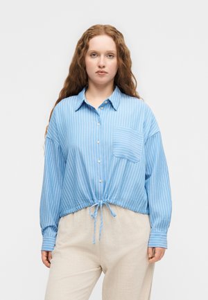 Γυναίκα με μακριά σγουρά μαλλιά που φοράει μπλε και λευκό ριγέ πουκάμισο με κουμπιά δεμένο στη μέση και μπεζ παντελόνι, στέκεται μπροστά σε ένα απλό φόντο.