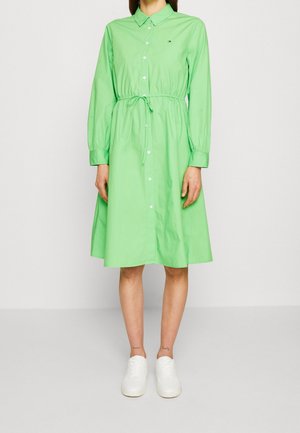 Frau in einem hellgrünen, knopfbaren Kleid mit langen Ärmeln, einem Taillengürtel und weißen Turnschuhen, die vor einem schlichten Hintergrund steht.