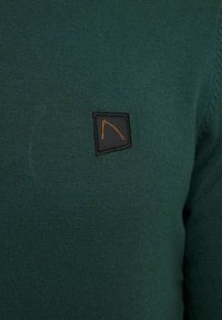Dunkelgrüner Strickpullover mit einem kleinen schwarzen und orangefarbenen Logopatch im linken Brustbereich. Glatte Textur mit einer taillierten Passform.
