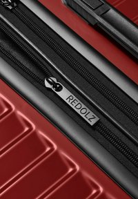 Primer plano de una maleta roja con cremallera negra que presenta un tirador de metal grabado con "REDOLZ" y un mango texturizado.