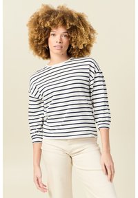 Gestreiftes Oberteil in Weiß und Marineblau mit Dreiviertelärmeln und Rundhalsausschnitt. Weicher, strukturierten Stoff. Das Modell trägt hellfarbene Hosen.