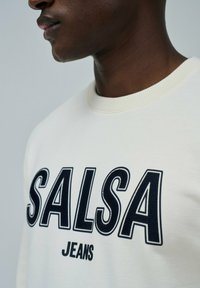 Sudadera crema con el texto en negrita en azul marino "SALSA" y "JEANS". Presenta un cuello acanalado y una textura suave. Primer plano del área del hombro y el cuello.