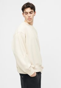 Νεαρός με σκούρα μαλλιά που φορεί εκρού φαρδιά φούτερ και μαύρο παντελόνι, με τα χέρια στις τσέπες, κοιτάζει προς τα κάτω αριστερά του.