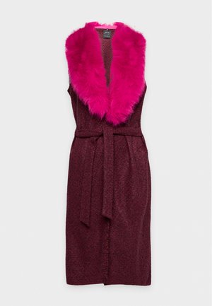 Une femme porte une longue jupe bordeaux et un manteau sans manches à col en fourrure rose vif, un chemisier crème et des bottines noires.