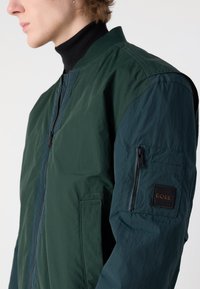 Jachetă bomber verde cu o textură ușoară, dotată cu fermoar frontal, guler ribbed și un buzunar cu fermoar pe mânecă stângă. Logo-ul brandului este vizibil.