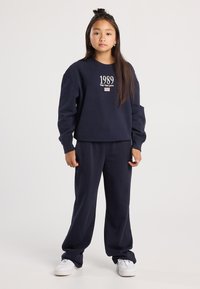 Niña joven de pie, vistiendo un sudadera azul marino con "1989 New York State" y pantalones a juego, zapatillas blancas, cabello largo, fondo neutro.