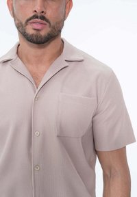 Chemise à manches courtes, couleur beige clair, en tissu à côtes texturé, avec un col noté et une poche poitrine unique avec fermeture à bouton.