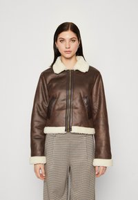 Giacca corta in pelle marrone con colletto e polsini in shearling bianco, dotata di una cerniera frontale e tasche laterali. Design strutturato e aderente.