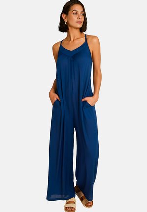 Femme aux cheveux courts et foncés portant une combinaison ample sans manches bleu marine avec des jambes larges et des poches, debout les mains dans les poches.
