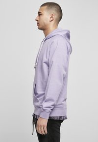 Sudadera de color púrpura claro hecha de material suave, con un bolsillo frontal, capucha con cordón y puños acanalados, llevada sobre una camisa de estampado.