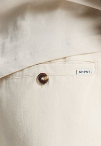 Light-colored fabric with a smooth texture, featuring a front pocket with a brown button and a white label reading "SHIWI."