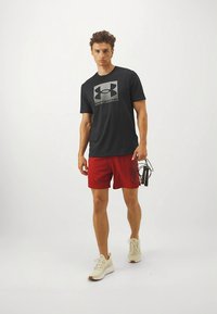 Melnas īsroku T-krekls ar pelēku Under Armour logotipu, kombinēts ar sarkaniem sporta šortiem. Modelis tur rokās lecamauklu, valkā vieglus sporta apavus.