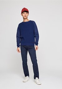 Pull en maille bleu marine avec une texture côtelée, porté avec un pantalon sombre et des baskets blanches, agrémenté d'un bonnet en maille rouge.