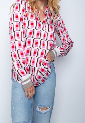 Femme portant un chemisier à manches longues à motif géométrique avec des carrés roses et rouges, associé à un jean bleu clair déchiré.