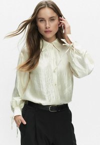 Crème gekleurde, lichte blouse met een knoopsluiting aan de voorkant, kraag en pofmouwen die bij de manchetten zijn gestrikt. Gecombineerd met zwarte broeken.