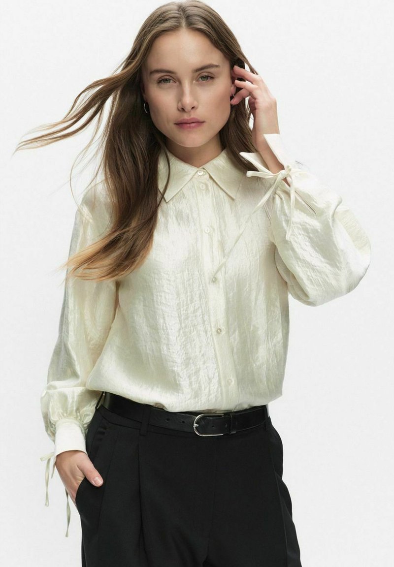 Crème gekleurde, lichte blouse met een knoopsluiting aan de voorkant, kraag en pofmouwen die bij de manchetten zijn gestrikt. Gecombineerd met zwarte broeken.