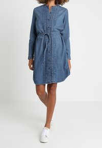 Jeansskjolklänning med knäppt front, långa ärmar, midjesnörning och rundad nederkant. Ljusblå färg med subtila vertikala sömmar.